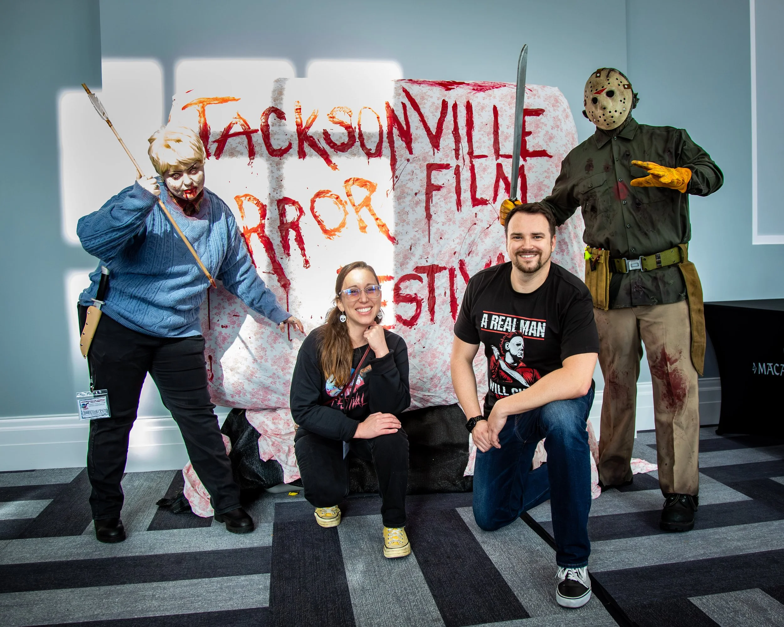 Two people dressed as horror movie killers with masks and costumes pose with two attendees at a horror film event. The backdrop features a large slime cover with red spray-painted lettering that reads "Jacksonville Horror" and "For" with additional w