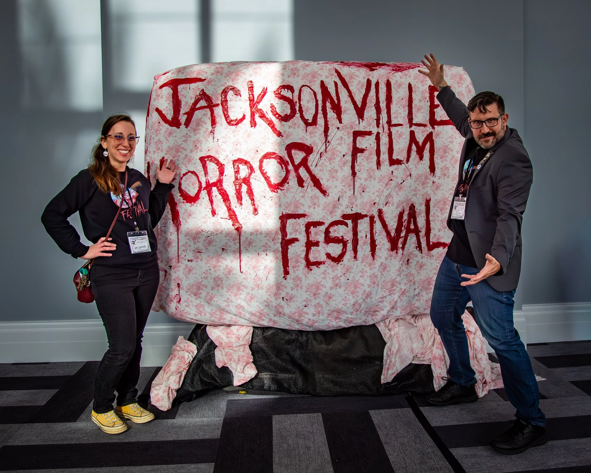 Two people standing next to a large pillow with the words 'Jacksonville Horror Film Festival' written in red, with one person pointing to it and the other smiling at the camera.