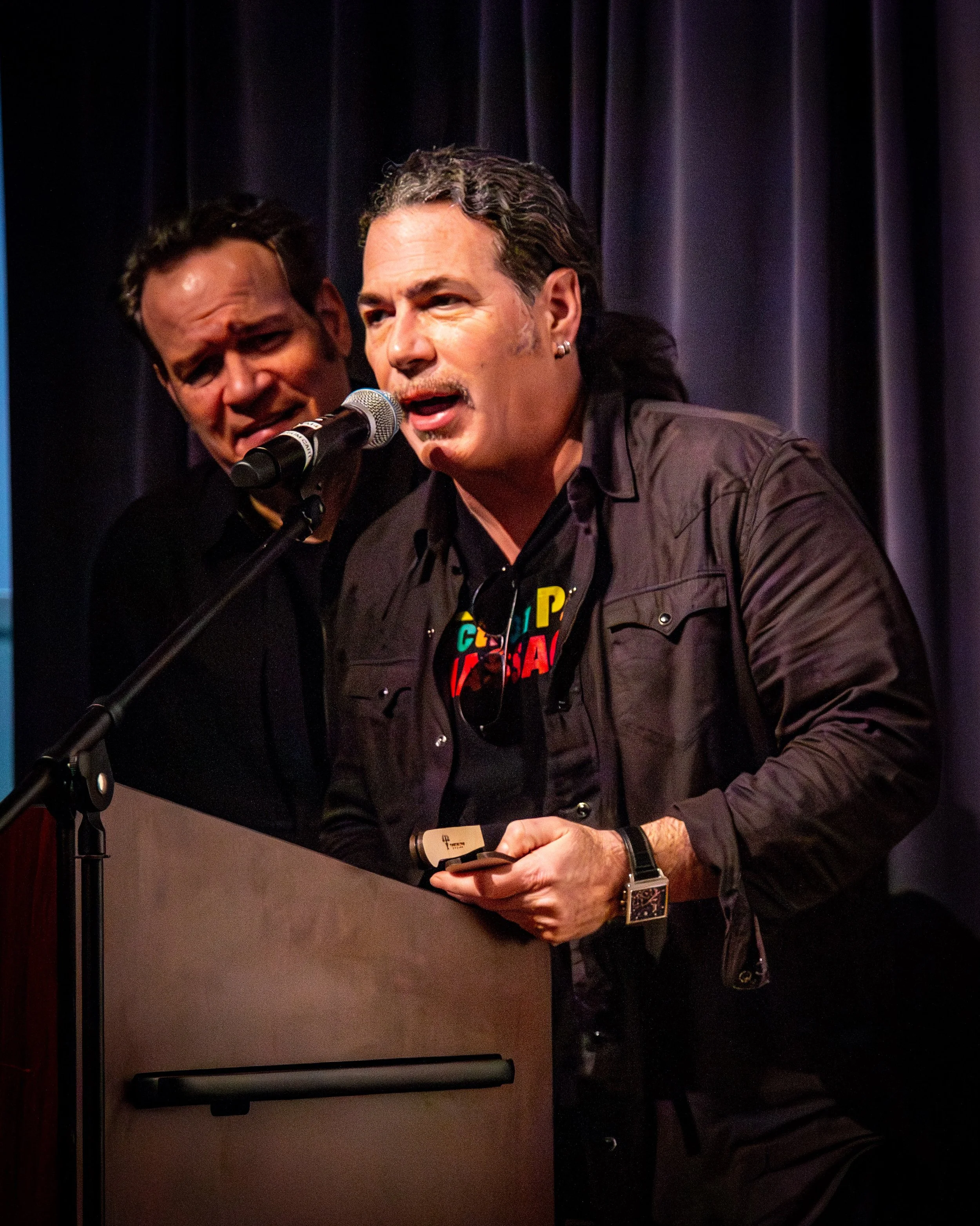 Two men speaking into a microphone on a stage, with blue curtains in the background. One man is wearing a black shirt with colorful text and a dark jacket, the other man behind him appears to be singing.