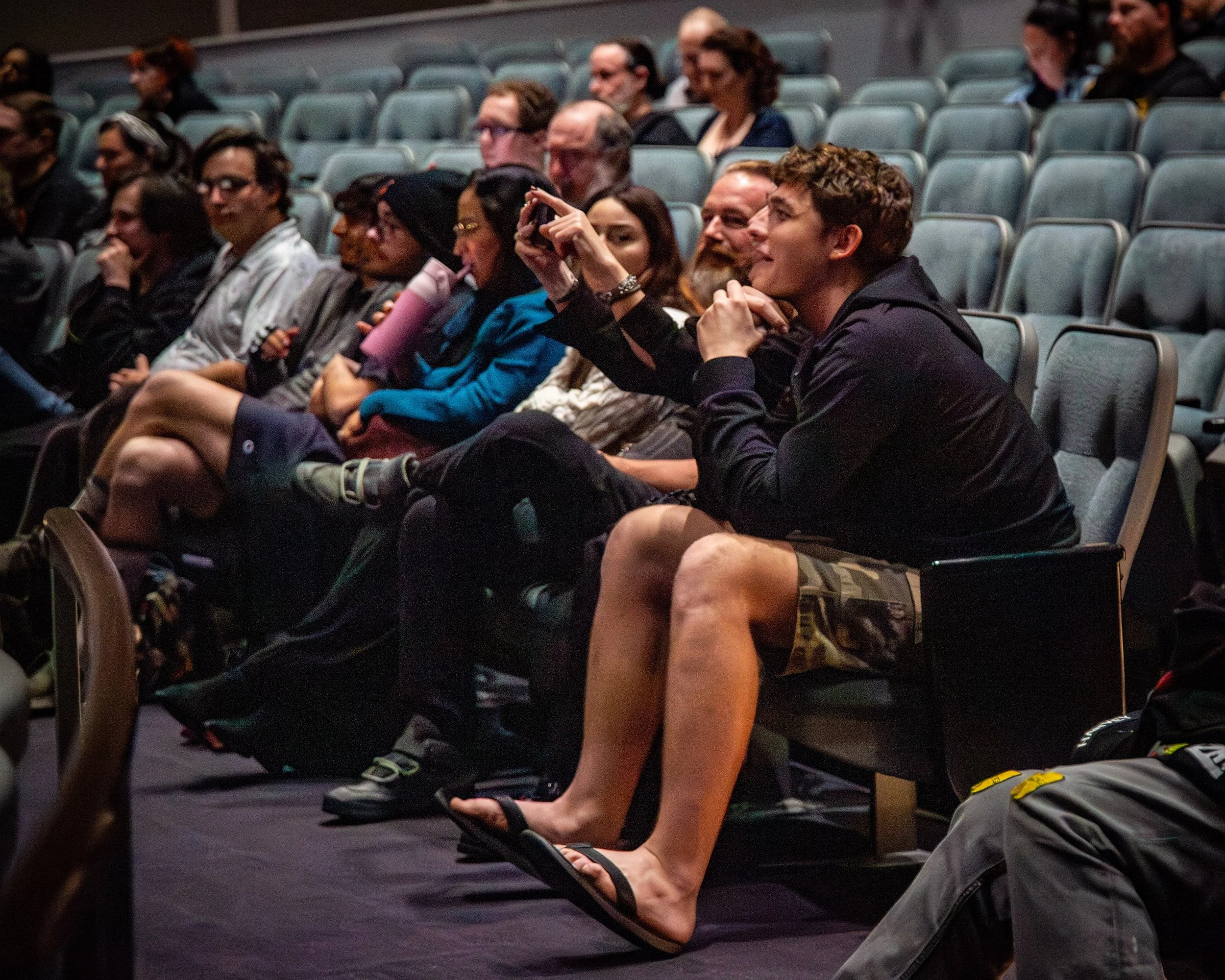Audience members seated in auditorium, some taking photos or watching, in a dimly lit room with gray seats.