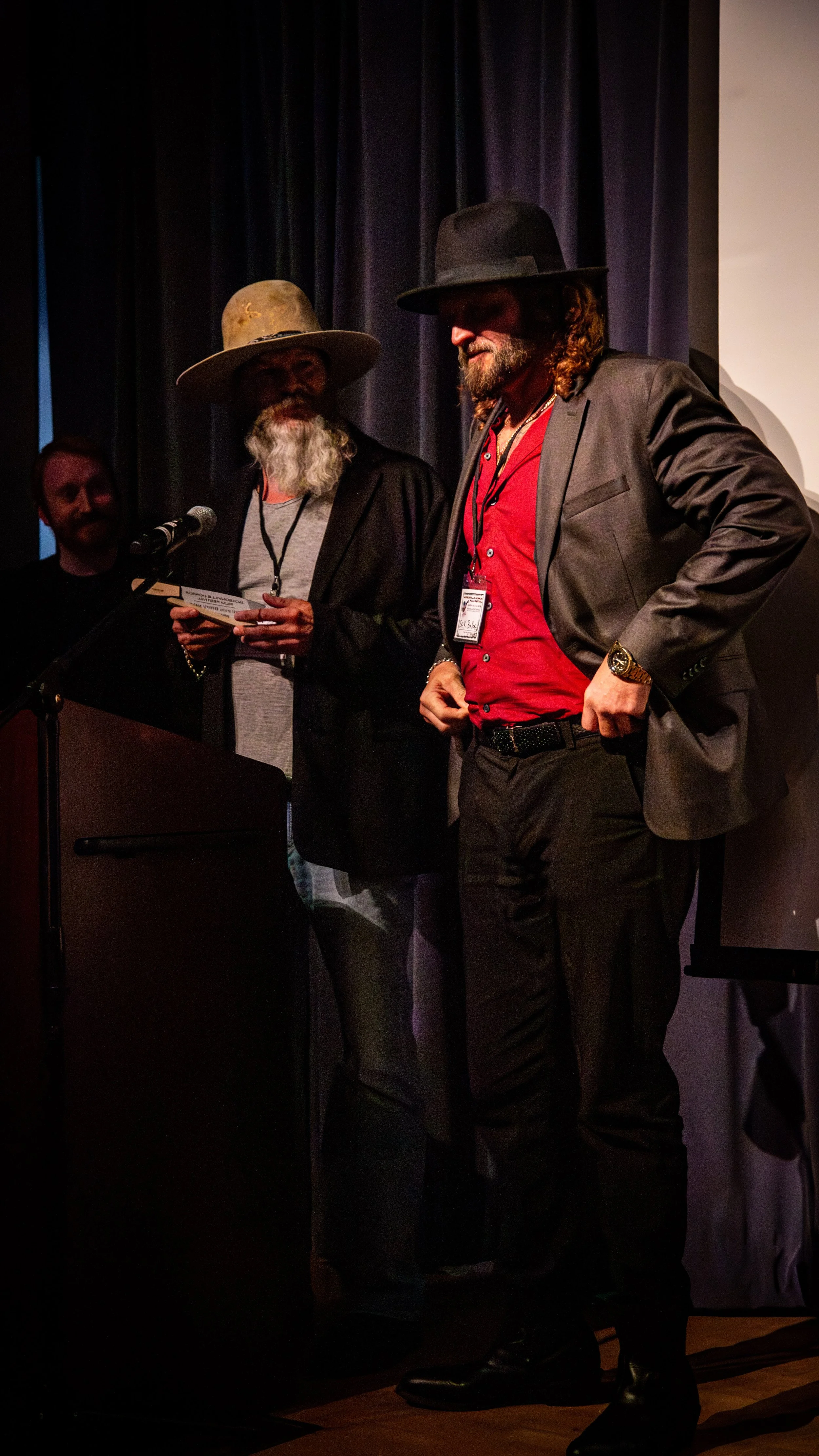 Two men standing on stage, one holding a paper, wearing hats and glasses, with a dark curtain in the background.