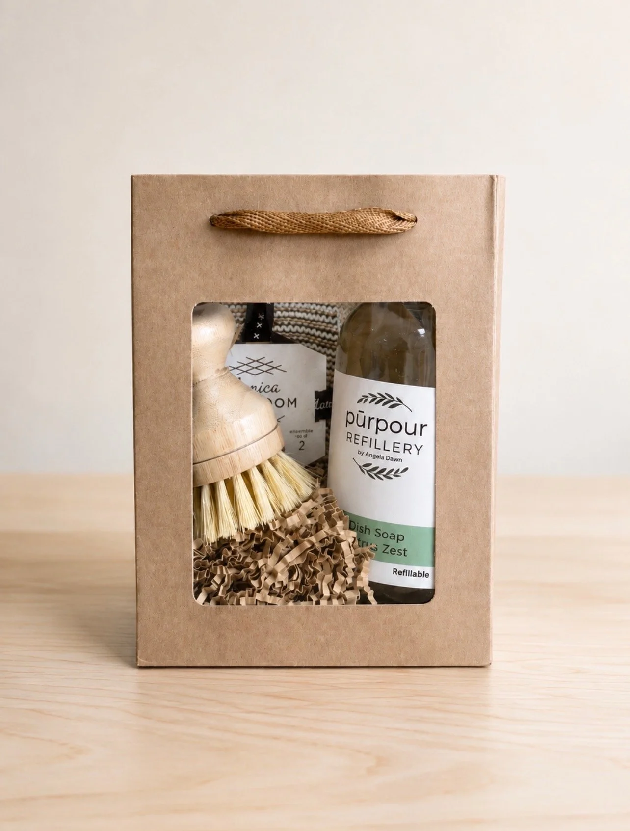 a paper gift bag with a clear window revealing a refillable bottle of dish soap, a dish scrubber know (with replaceable scrubbing knob) and a set of 2 dish cloths