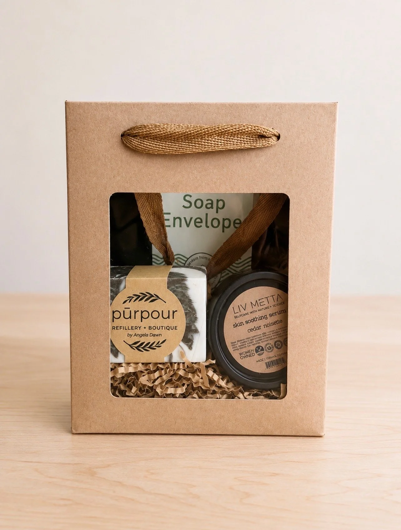 a paper gift bag with a clear window revealing a bar of natural soap, a jar of Liv Metta Cedar Noir Skin Smoothing Sugar Scrub, and a Soap Envelope