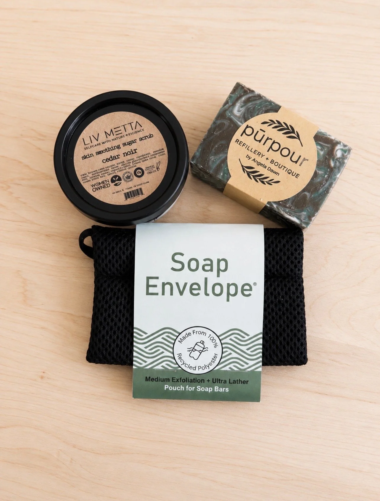a flatlay image of a bar of natural soap, a jar of Liv Metta Cedar Noir Skin Smoothing Sugar Scrub and a black mesh soap envelope