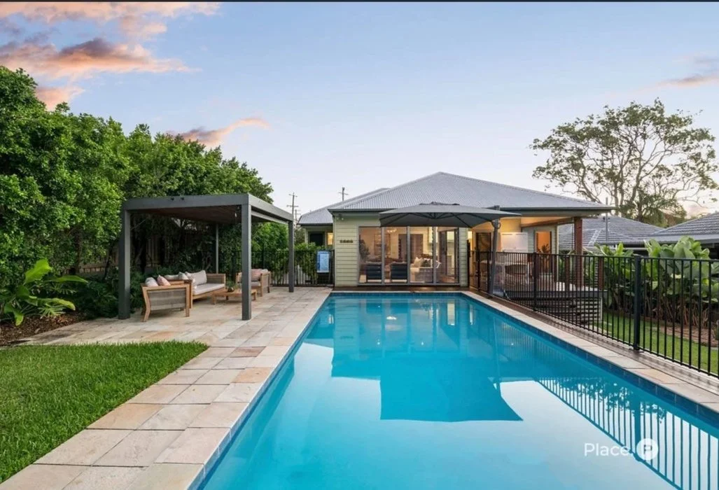 Backyard swimming pool with a patio and seating area, surrounded by green trees and a fence, house with large windows in the background, during sunset.