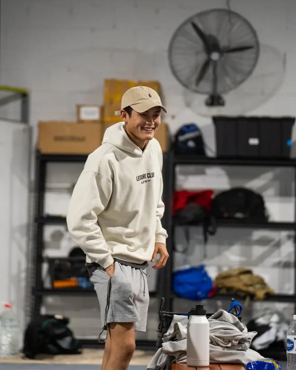 A young man dressed in a beige hoodie, gray shorts, and a beige cap, smiling and standing in a room with shelves and supplies, possibly a gym or storage area.