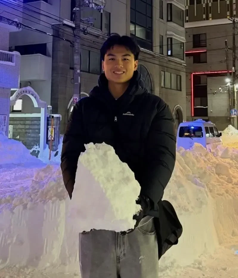 Smiling young man in a black jacket holding a large block of snow, standing outdoors at night on a snowy city street with buildings and lights in the background.