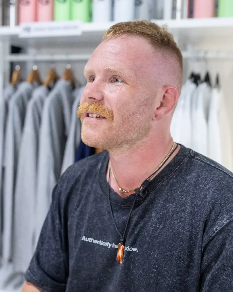 A man with red hair, a beard and Mustache, wearing a black t-shirt and a necklace, standing near clothing racks in a store or boutique.