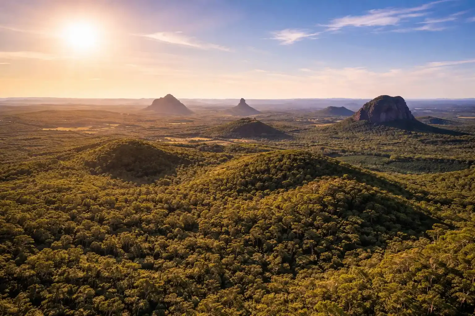 Aerial landscape of the Glass House Mountains in Queensland, showing lush green hinterland forests and volcanic peaks under a golden sunset sky.
