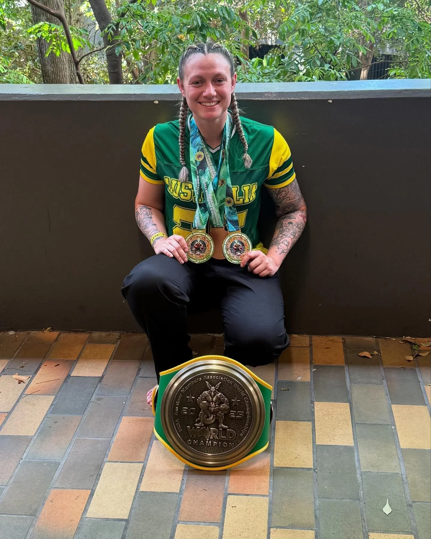 Person kneeling outdoors wearing a green and yellow jersey, holding two medals with a large world champion belt displayed in front of them.