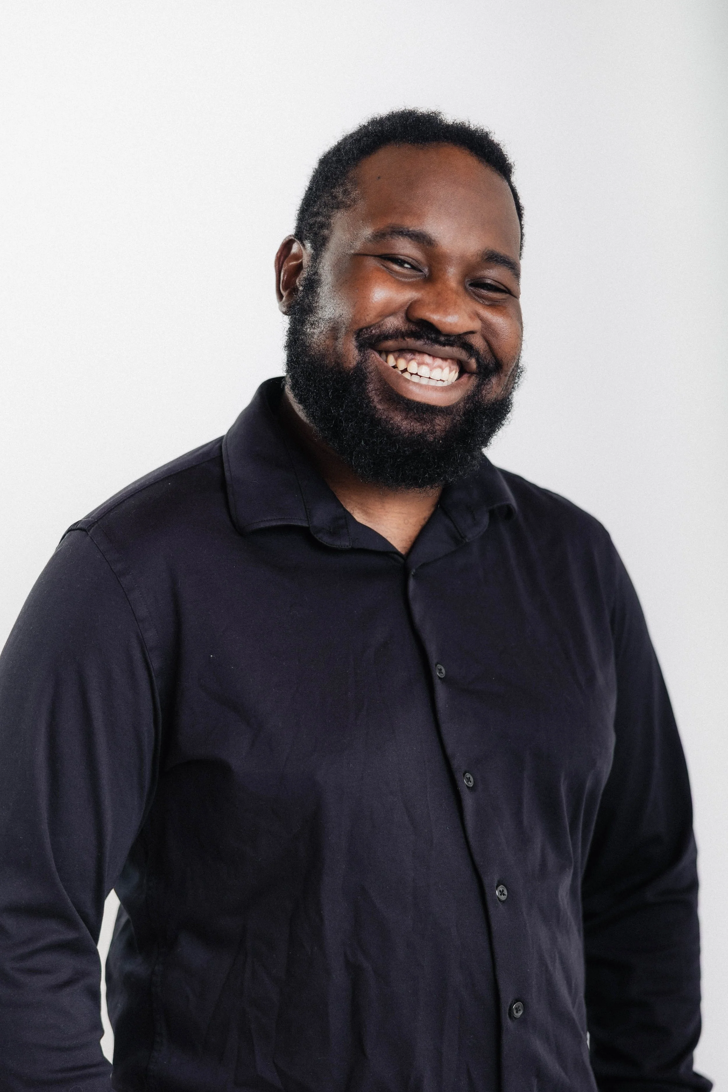 A smiling man with a beard and short black hair, wearing a black button-up shirt, standing against a plain white background.