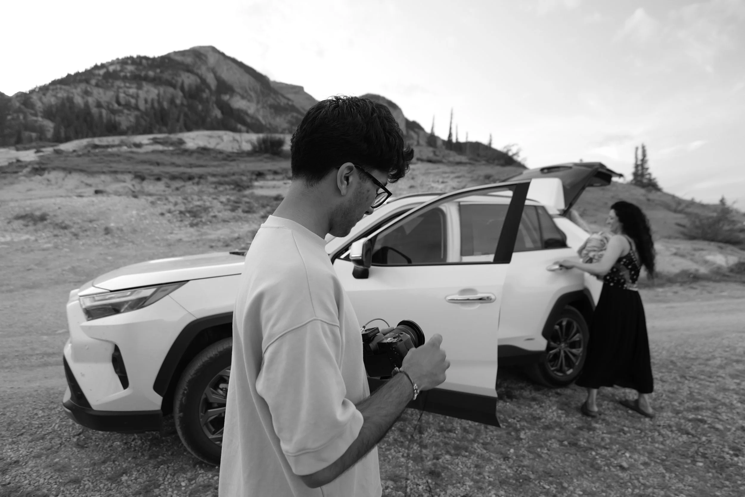 A man with glasses looking at a camera in a scenic outdoor setting with a woman near an open car hatchback.