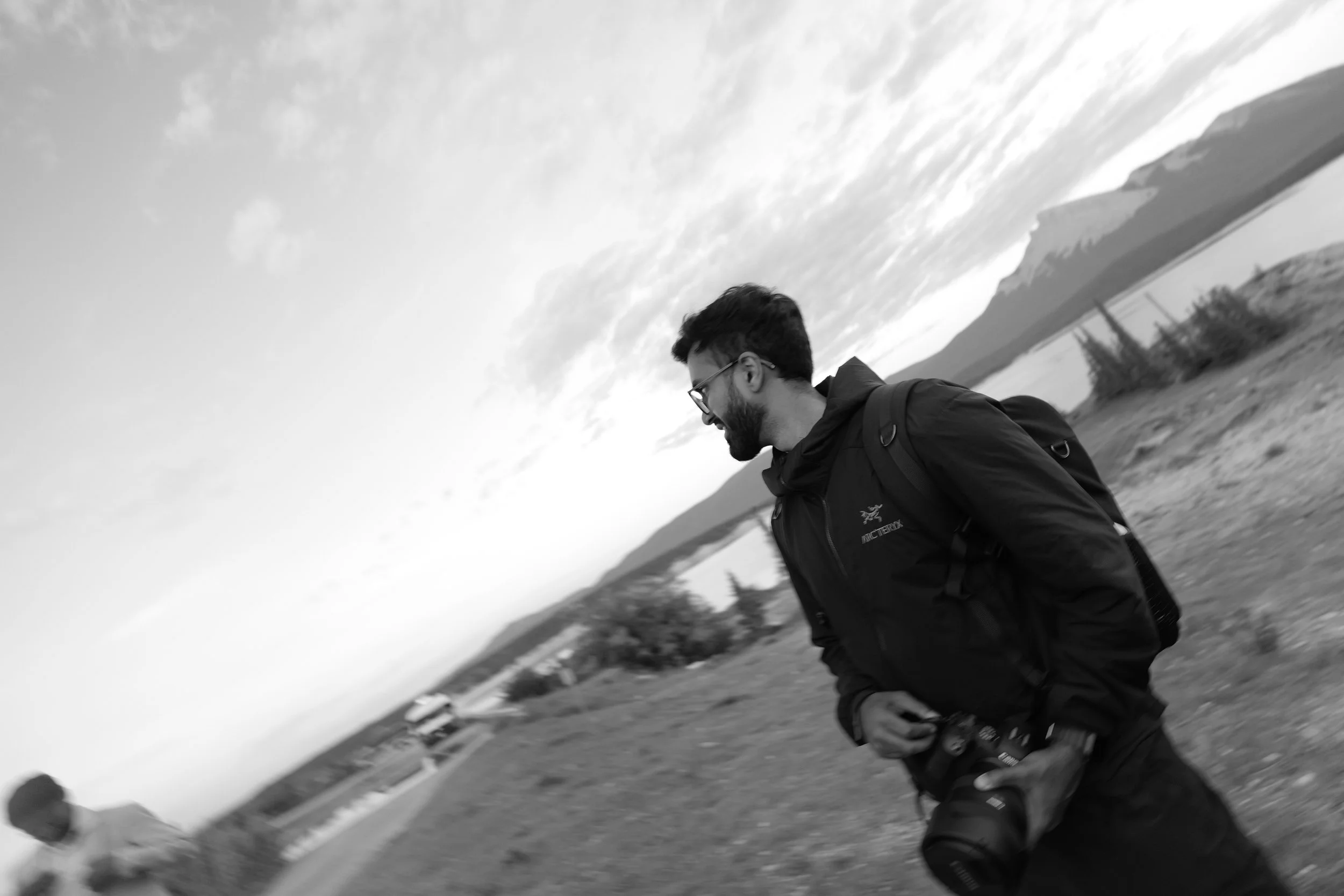 A man with glasses and a beard dressed in outdoor clothing, holding a camera, standing on a grassy area near a lake or river with mountains in the background, under a cloudy sky with some sun.