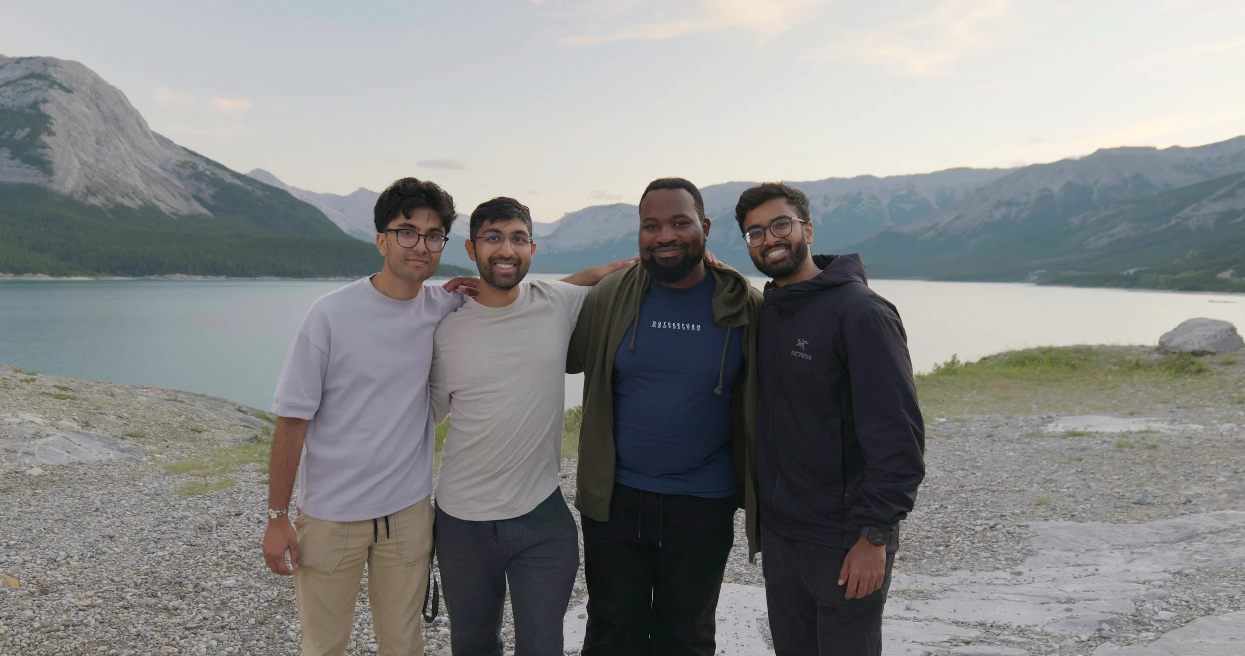 Four men standing together outdoors near a lake with mountains in the background, smiling, with their arms around each other.