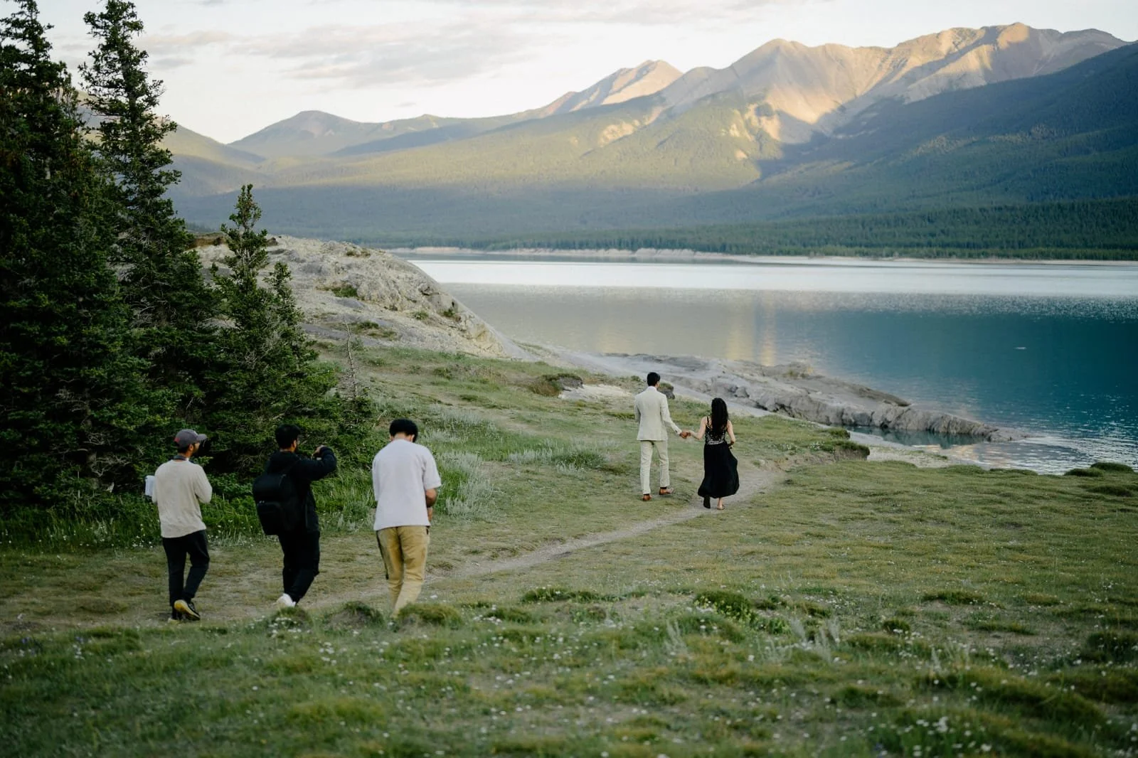 A group of five people walking on a grassy path near a lake with rocky shores, surrounded by trees and mountains in the distance during daylight.