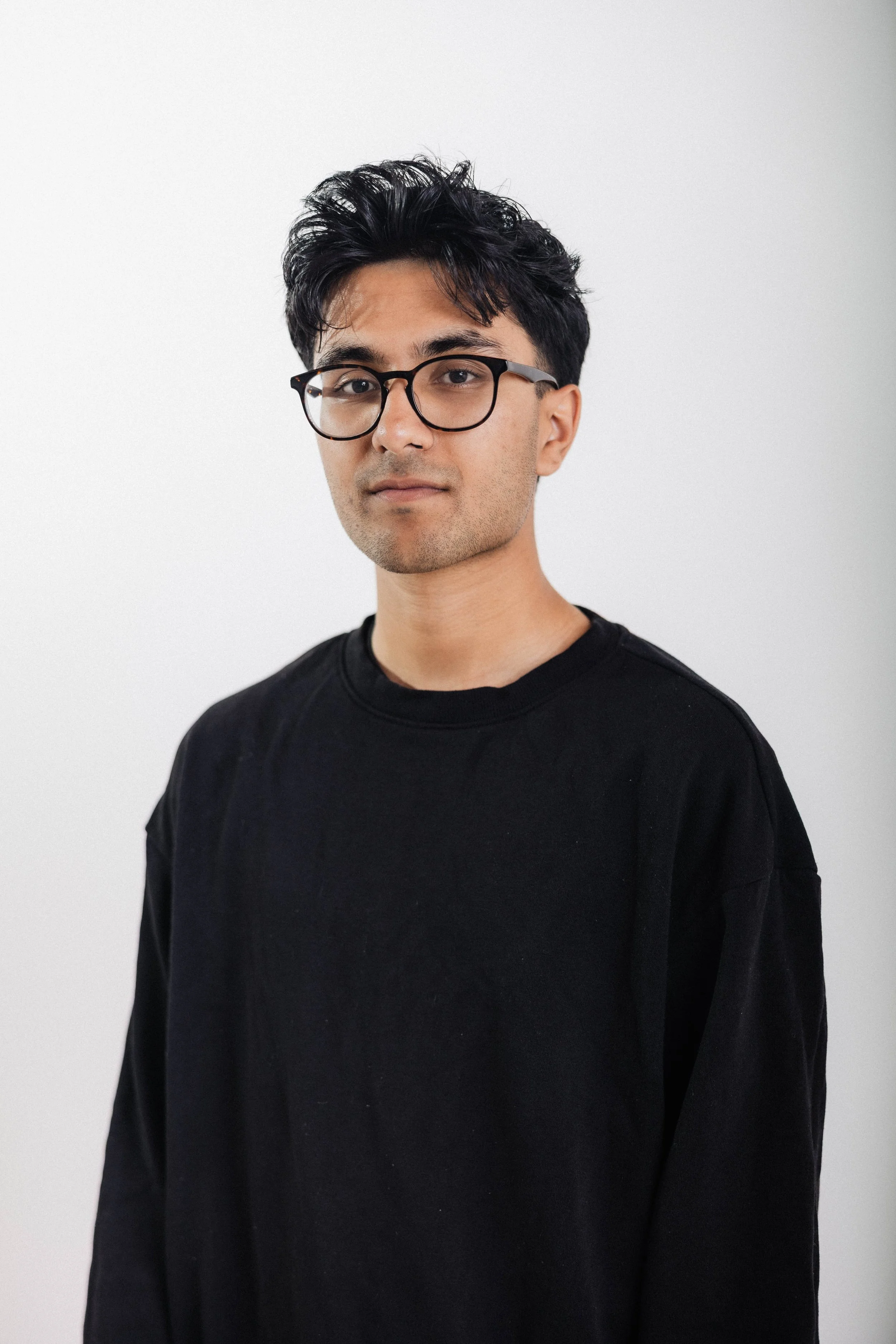 A young man with black hair, wearing glasses and a black shirt, standing against a plain white background.