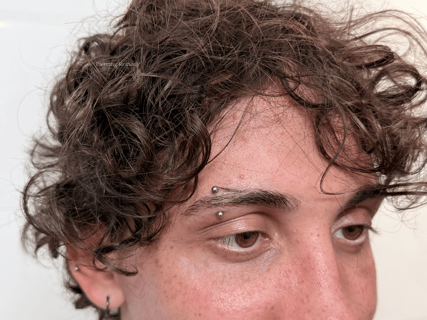 Close-up of a person's forehead and eyes, with dark curly hair and facial piercings above the eyebrow and on the earlobe.
