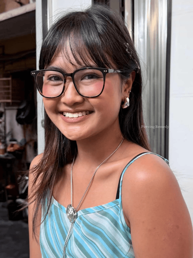 A young woman with medium skin, dark hair, glasses, and jewelry smiling outdoors in front of a metallic and wooden structure, wearing a strappy blue patterned top.