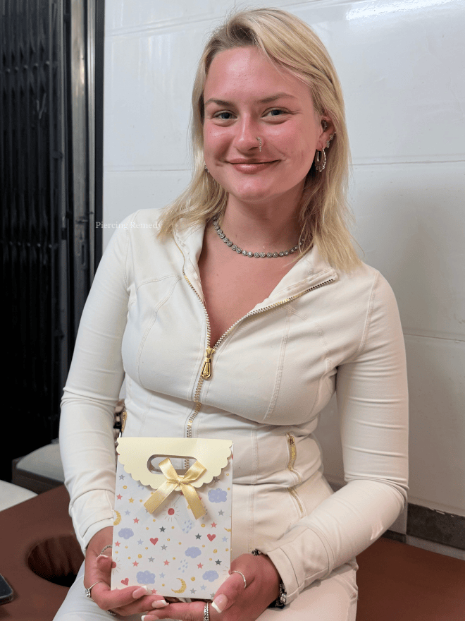 A young woman with blonde hair, wearing a white zip-up jacket, jewelry, and earrings, smiling and holding a decorated gift bag with a bow.