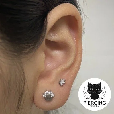 Close-up of a person's ear with three silver earrings, including a cubic zirconia and a small black circle, with a logo reading 'Piercing Remedy' featuring a black cat.