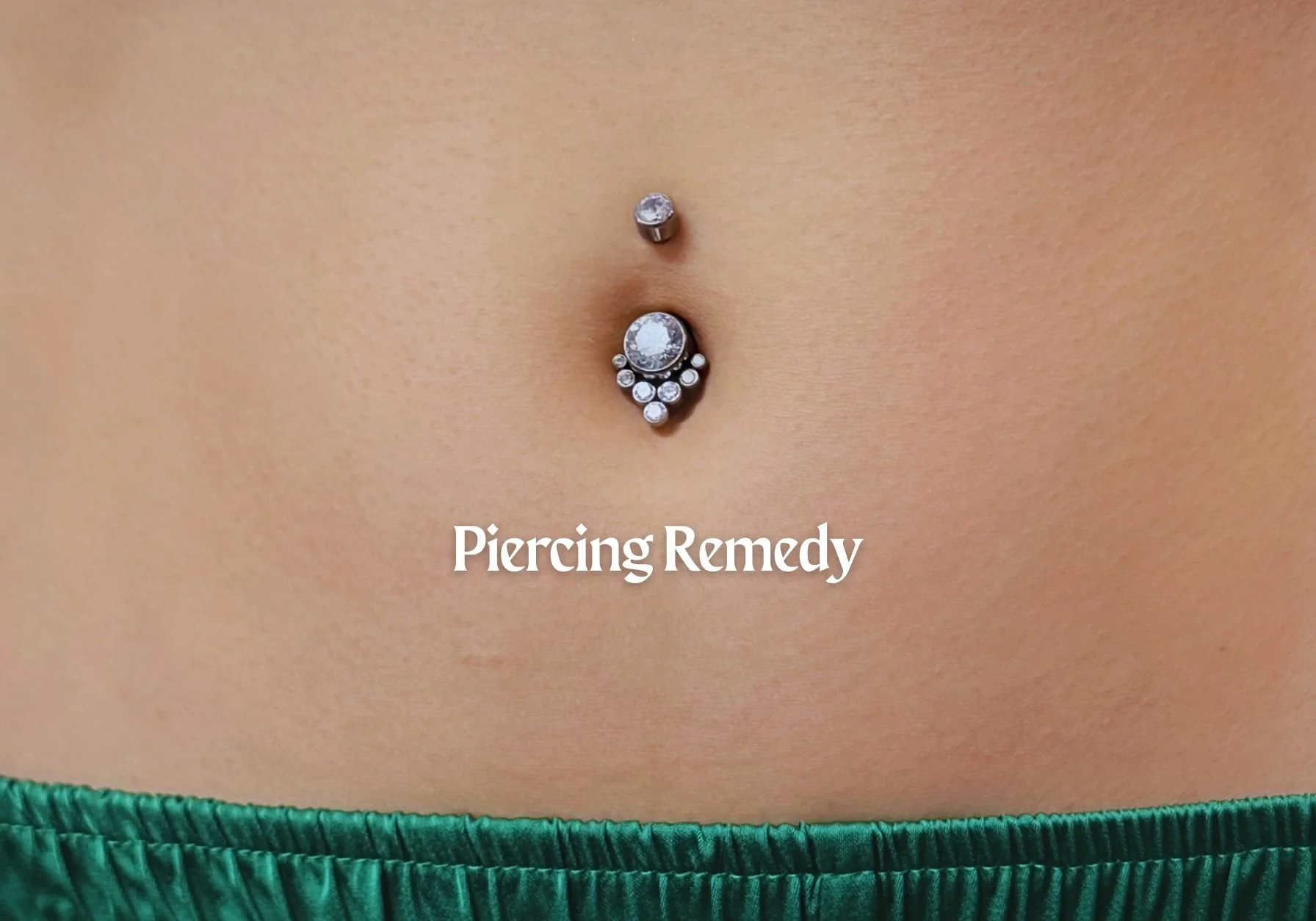 Close-up of a person's belly with a navel piercing adorned with several rhinestones, against a background of green fabric. Text on the image reads 'Piercing Remedy'.