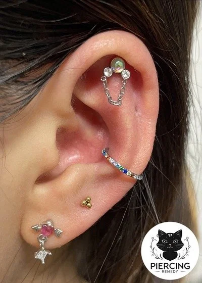 Close-up of multiple earrings on a person's ear, including a cartilage chain, a hoop with multicolored stones, a small gold stud, and a pink and silver pendant earring with a wing and star design.