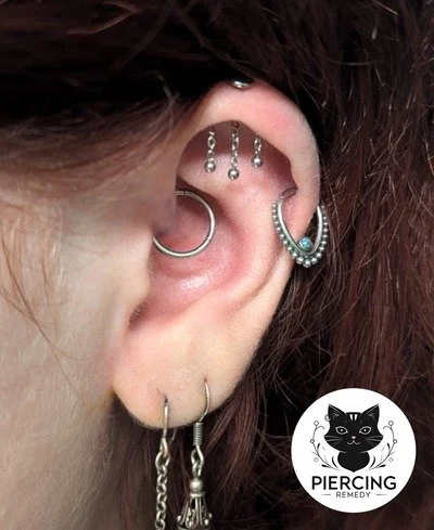 Close-up of a person's ear with multiple silver earrings, including a hoop, a cuff, and dangling chains, and a black cat logo with the text "Piercing Remedy".