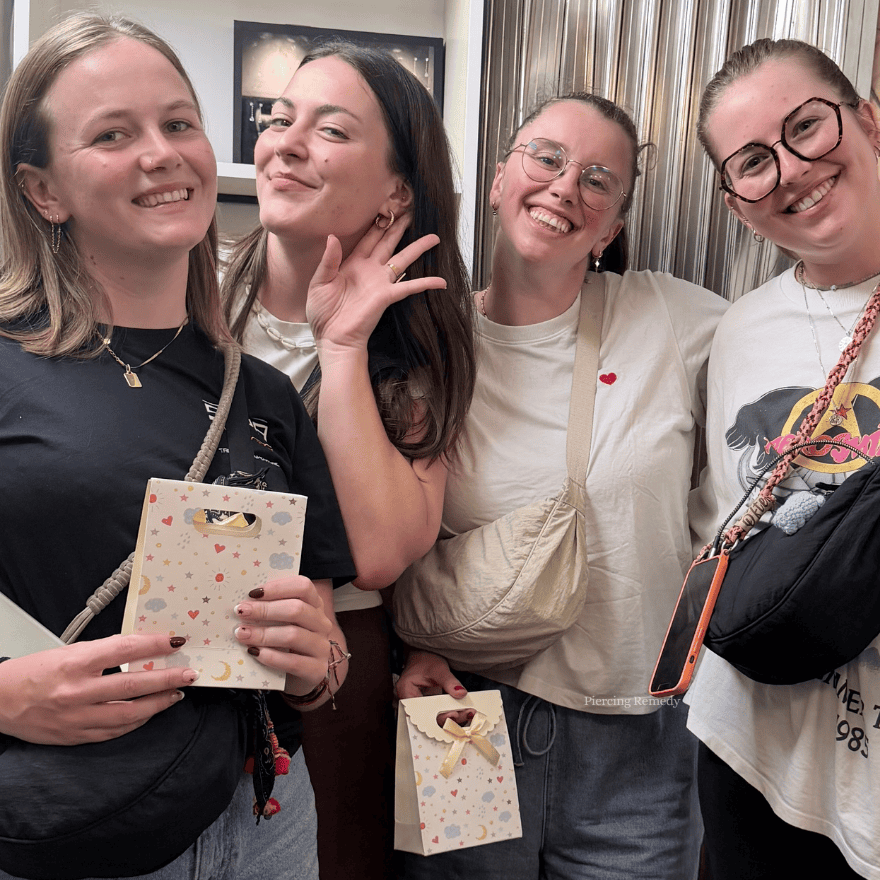Four women standing together indoors, smiling at the camera, holding small gift bags with star and moon patterns, and one wearing glasses with a necklace, casual clothes, and accessories.