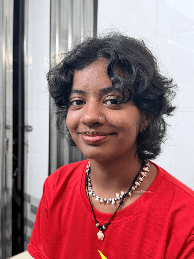 Young woman with short, curly black hair, wearing a red shirt and layered necklaces, smiling in an indoor setting with metal and glass background.