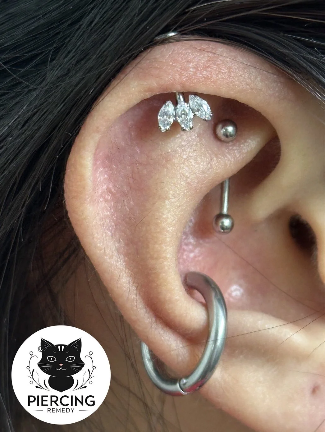 Close-up of a person's ear with multiple piercings, including a decorative diamond butterfly stud, a small metal ball stud, and a larger hoop earring, with dark hair partially covering the ear.