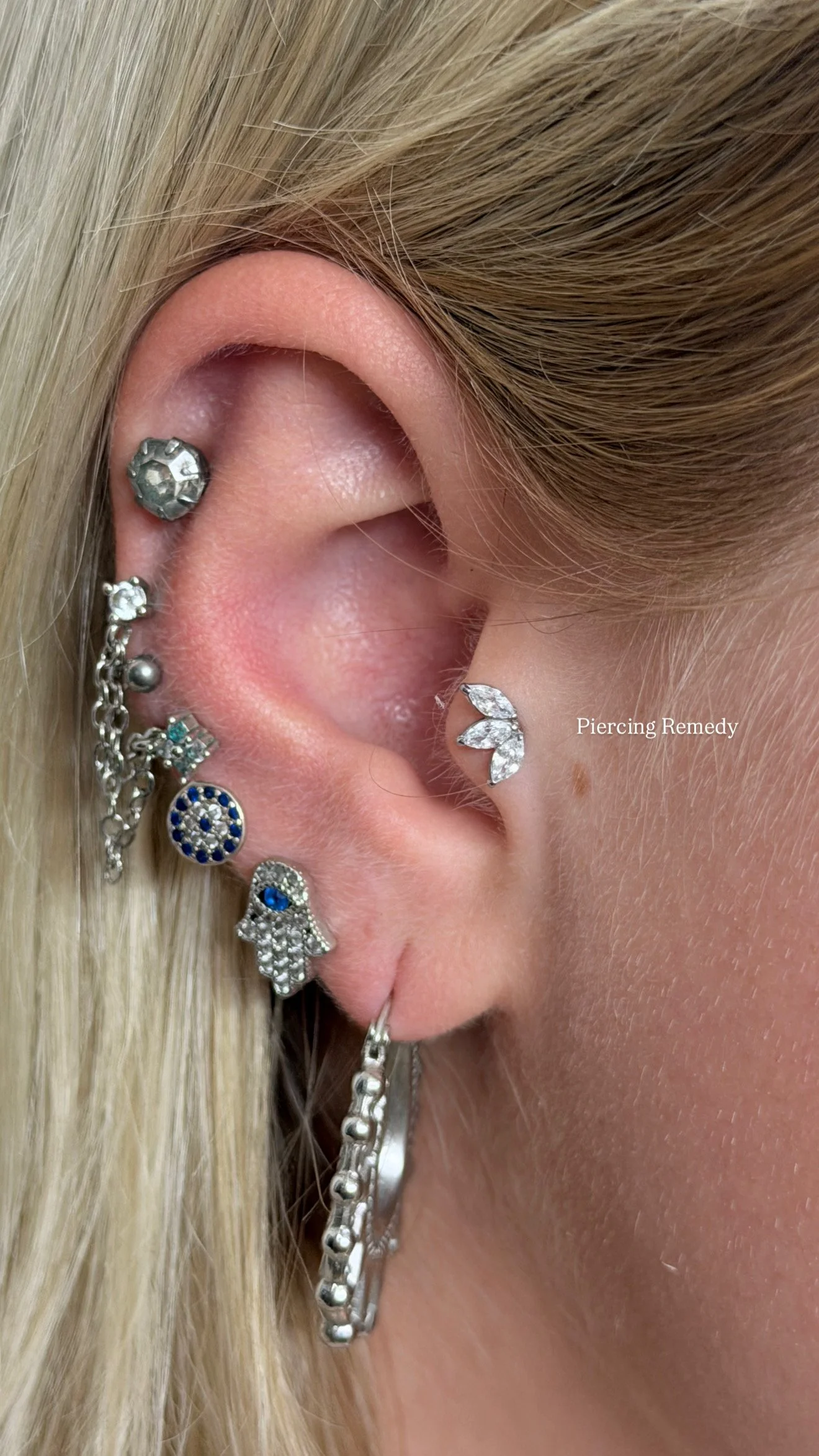 Close-up of a human ear with multiple piercings, including a cartilage piercing with a gem ring, a lobe piercing with a leaf-shaped diamond earring, and a hoop earring with beads. The skin appears healthy, and blonde hair partially covers the ear.