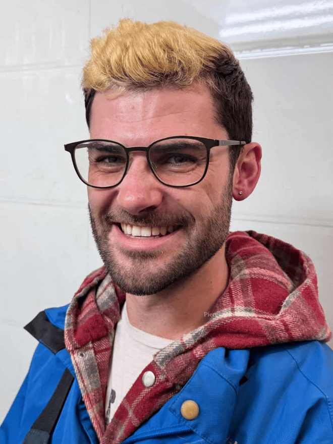 A smiling man with glasses and a beard, wearing a blue jacket over a red plaid hoodie, standing indoors.
