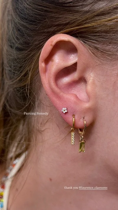 Close-up of a woman's ear with multiple earrings, including a small flower-shaped stud, a vertical bar earring, and a dangling rectangular gem earring.