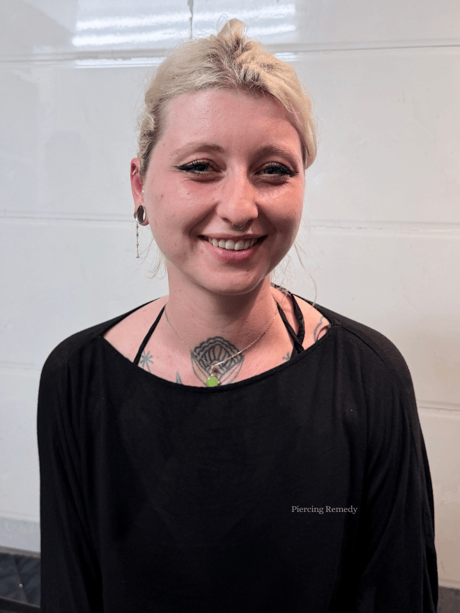 Smiling young woman with short blonde hair, tattoos on her chest, earrings, and wearing a black top, standing against a white wall.