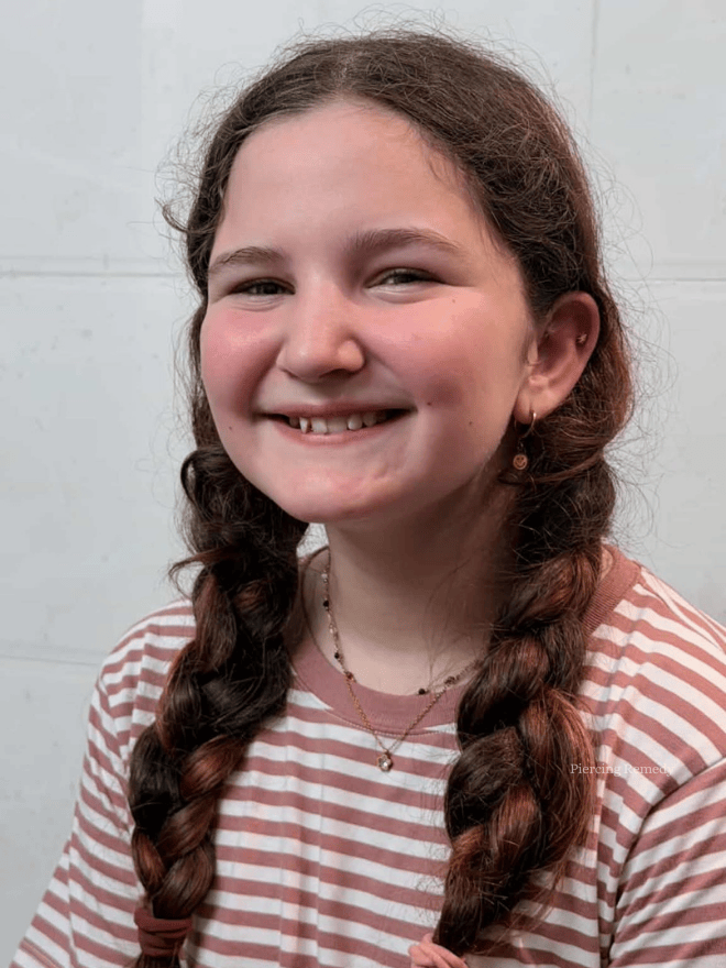 A young girl with long brown braided hair, smiling, wearing a striped pink and white shirt, gold earrings, and layered necklaces, standing against a light-colored wall.
