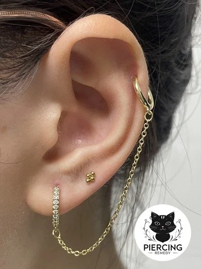 Close-up of a woman's ear with multiple piercings featuring gold and diamond jewelry, including a chain connecting two earrings, and a logo of a black cat with the words "Piercing Remedy."