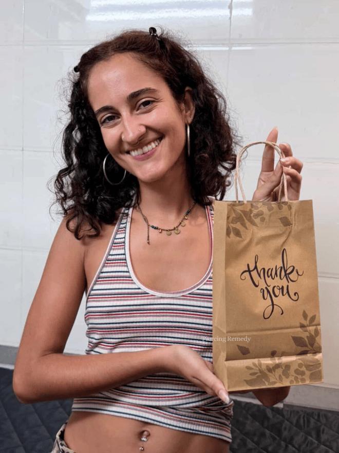 A young woman with curly brown hair, wearing hoop earrings and a striped tank top, smiling and holding a brown paper bag with "thank you" written on it.