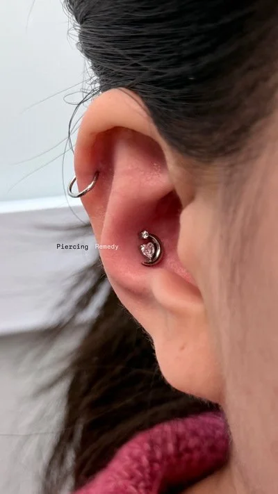 Close-up of a person's ear, showing two earrings: a small hoop earring in the upper cartilage and a moon-shaped earring with a small gem in the lower lobe.