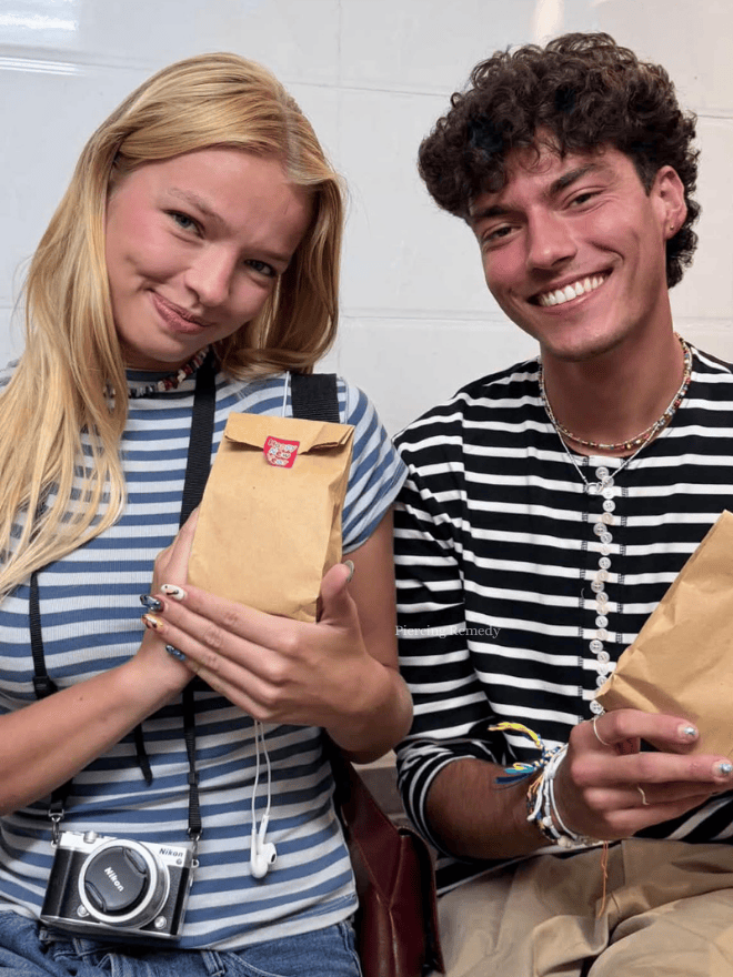 Two young people, a woman with long blonde hair and a man with curly brown hair, smiling while holding paper bags in an indoor setting.