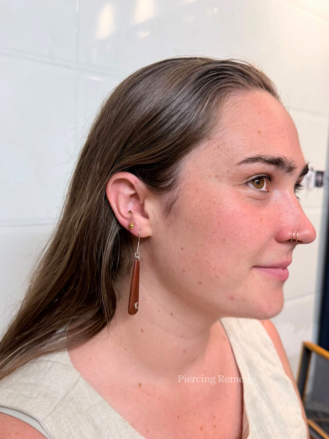 Side profile of a woman with long brown hair, wearing earrings and a nose ring, in front of a white background.