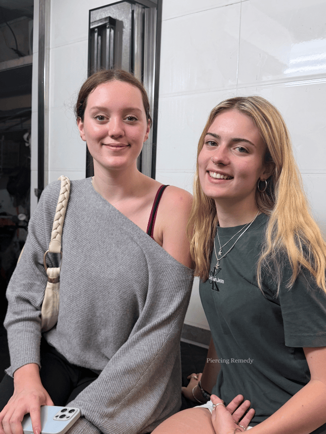 Two young women sitting close together, smiling, indoors with a white tiled wall behind them.