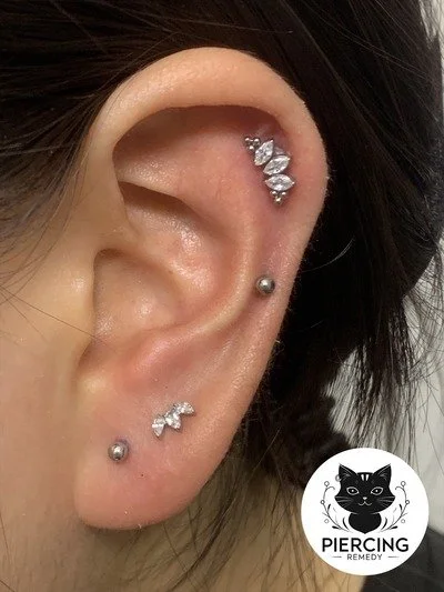 Close-up of a person's ear with multiple piercings, including a cartilage piercing with three small diamonds, a helix piercing with three small diamonds, a conch piercing with a small diamond, and two lobe piercings with metallic balls, along with a 