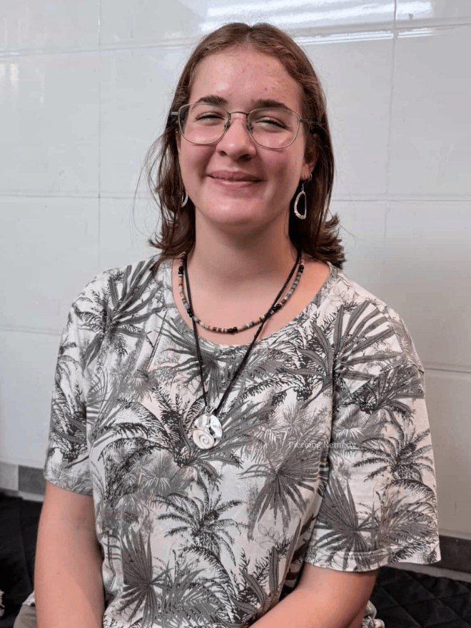 A young woman with glasses and earrings smiling at the camera, wearing a tropical print shirt and layered necklaces, standing against a tiled wall.