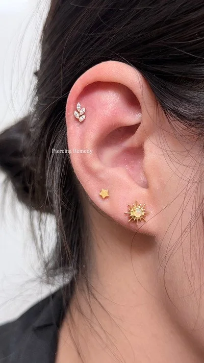 Close-up of a person's ear with three earrings: a silver butterfly-shaped earring at the top, a small gold star-shaped earring in the middle, and a gold sun-shaped earring at the bottom.