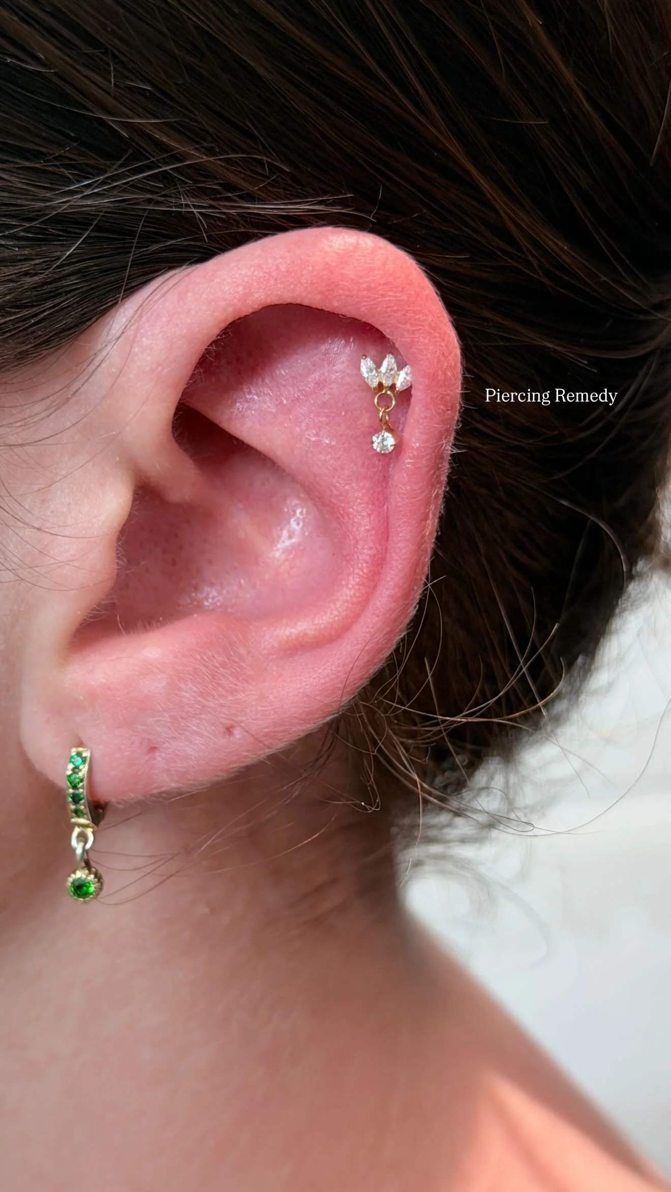 Close-up of a person's ear with two earrings: a small hoop with green stones on the earlobe and a dangle stud with clear stones in the cartilage. Text overlay says 'Piercing Remedy'.
