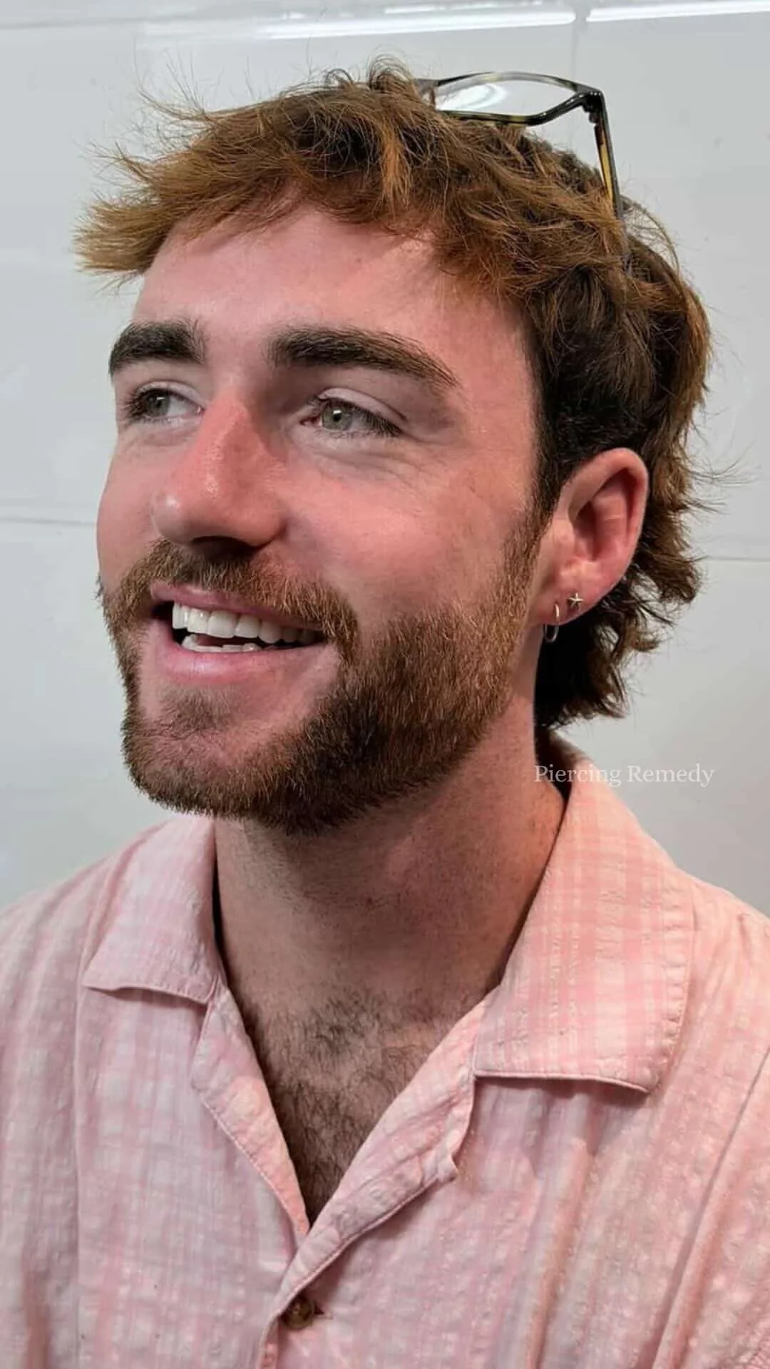 A smiling man with light-colored hair, a beard, and light gray eyes, wearing a pink checkered shirt and glasses on his head, in front of a white background.
