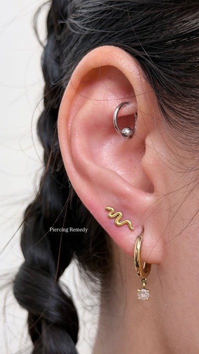 Close-up of a person's ear with multiple earrings, including a silver hoop with a ball, a gold snake-shaped earring, a small gold hoop, and a dangling earring with a clear gemstone. The person has dark hair styled in a braid.