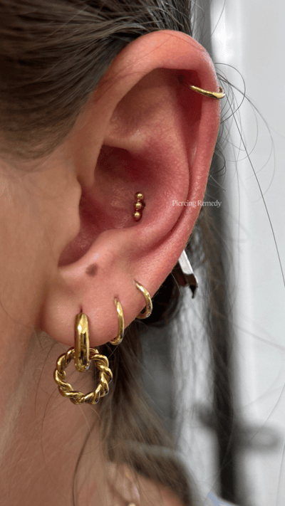 Close-up of a person's right ear showing multiple gold earrings, including a helix piercing, a cartilage piercing, a snug or daith piercing with gold ball studs, and various hoop earrings.