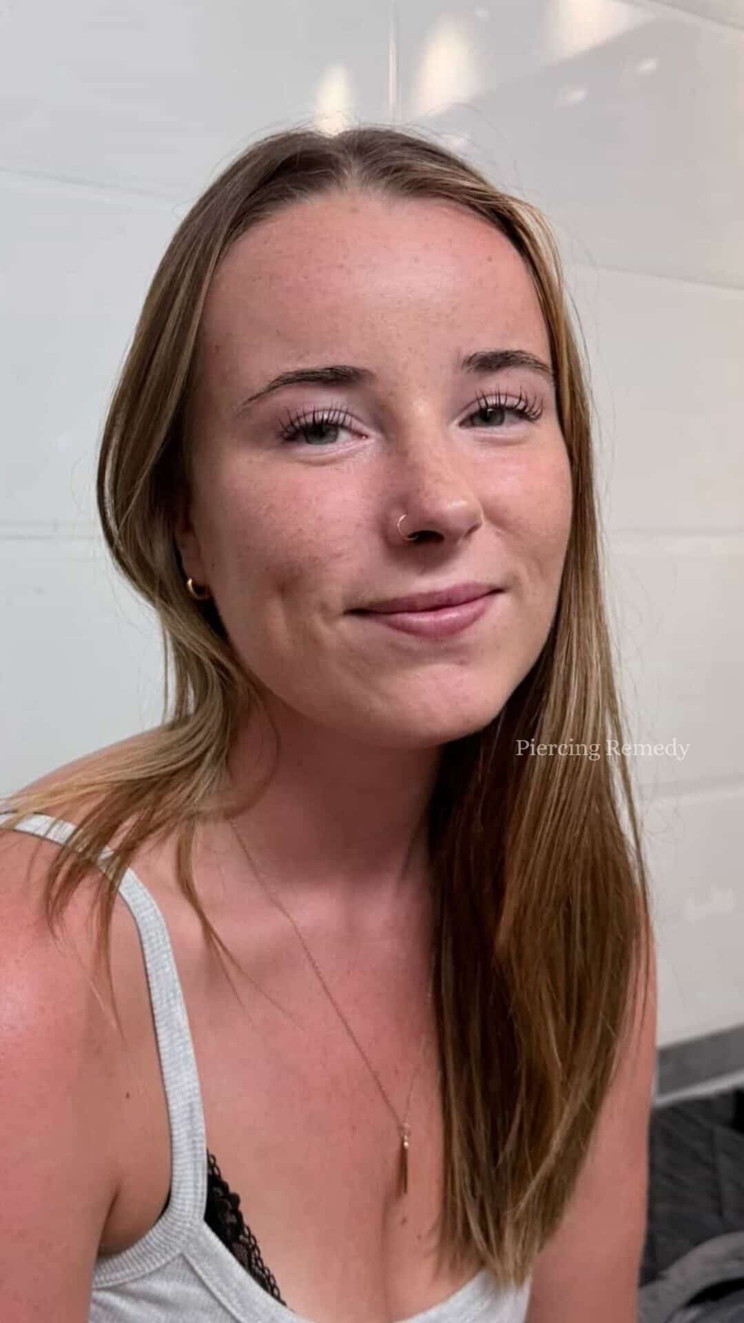 A young woman with long reddish-brown hair, wearing a white tank top with black lace trim, small hoop earrings, a nose ring, and a delicate necklace, smiling softly in front of a plain light-colored wall.