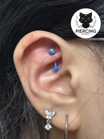 Close-up of a person's ear with a blue and silver helix piercing, a daisy flower earring, and a small silver hoop earring. A piercing logo with a black cat is in the top right corner.